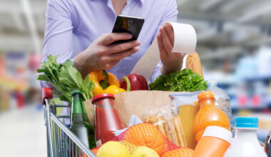 Person pushing a supermarket car full of groceries and looking at receipt and phone