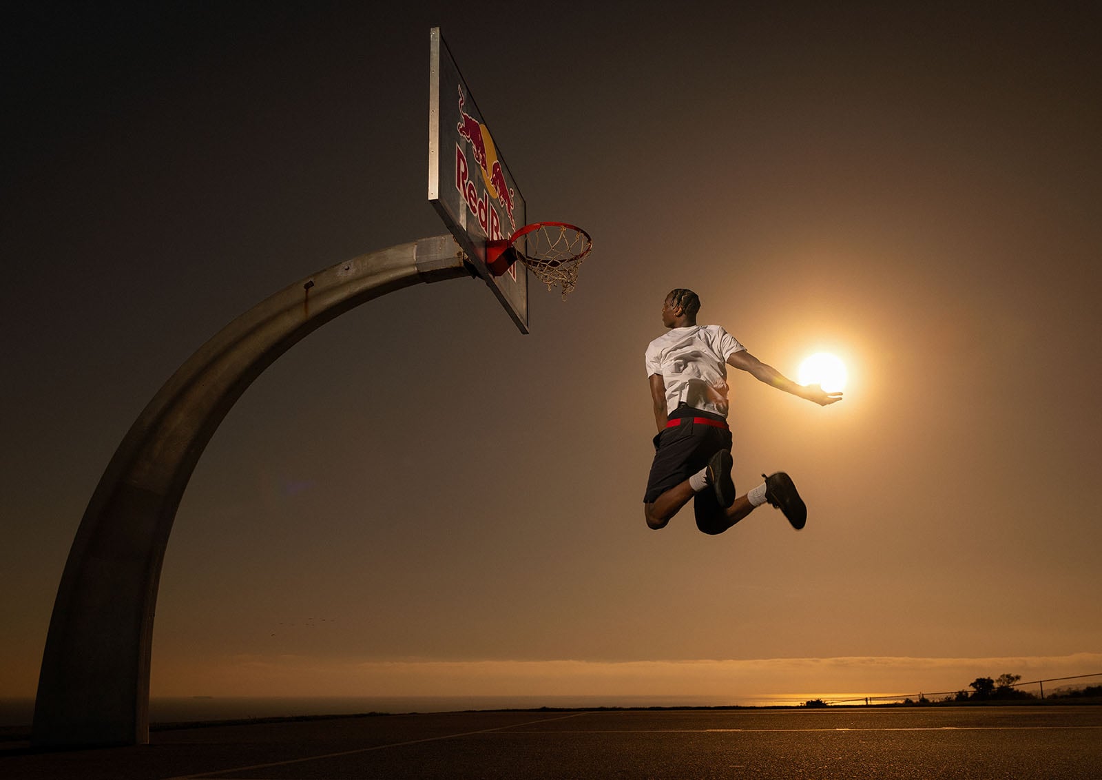 A basketball player leaps towards a hoop at sunset, holding his arm out so the setting sun appears like a glowing basketball in his hand. The sky is golden, and the outdoor court is silhouetted in the warm light.