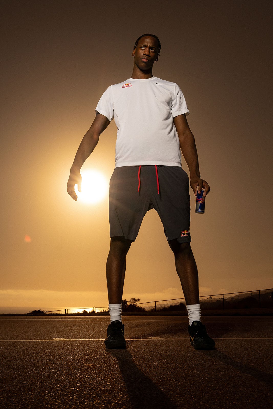 A man stands outdoors at sunset, wearing a white T-shirt and dark shorts with red drawstrings. He holds a can in his right hand, with the sun low in the sky behind him and a fence in the distance.