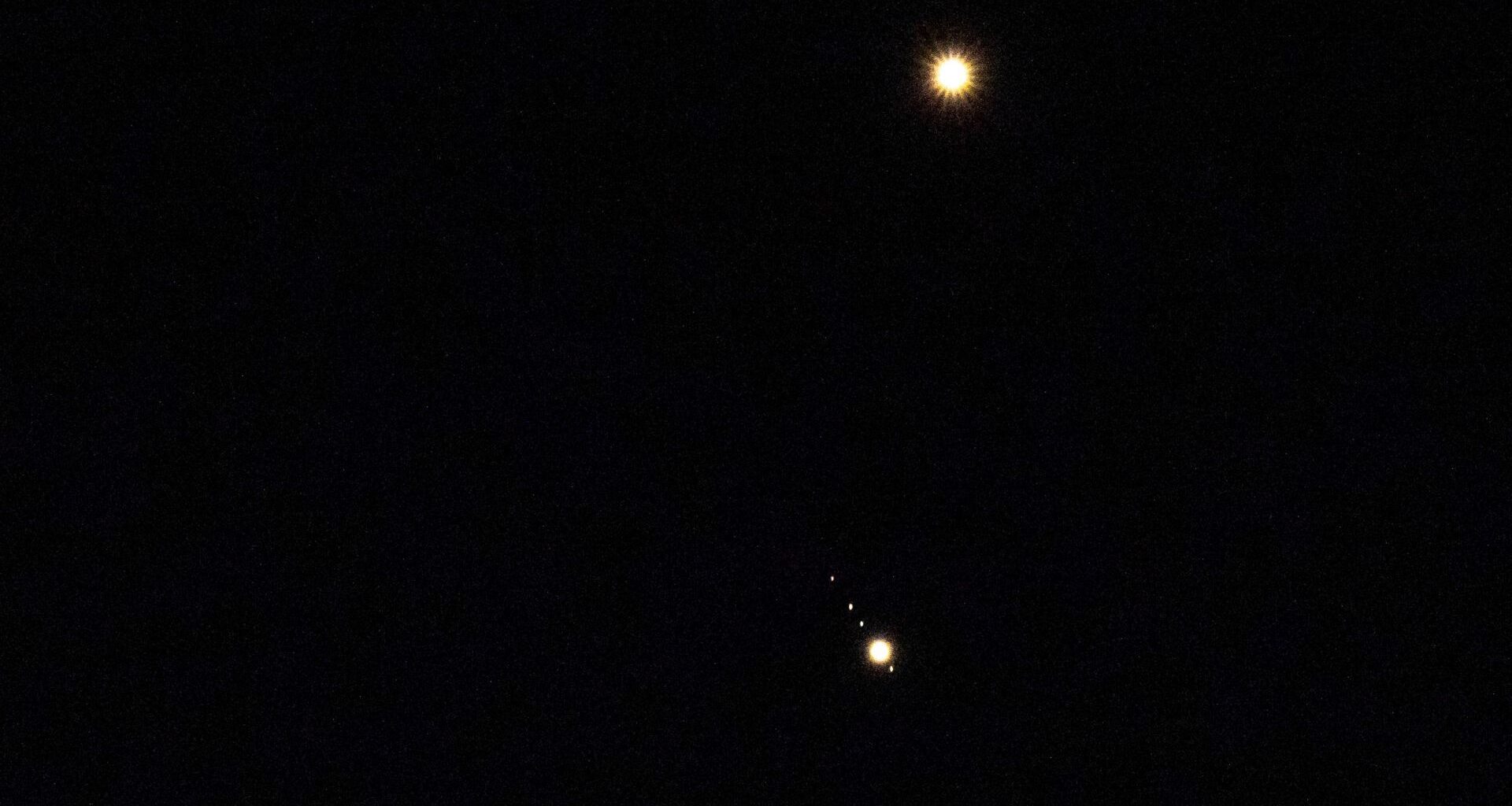 Venus is pictured shining brightly above Jupiter in a black sky. A number of small star-like objects can be seen in a line surrounding Jupiter, representing its four largest Galilean Moons.