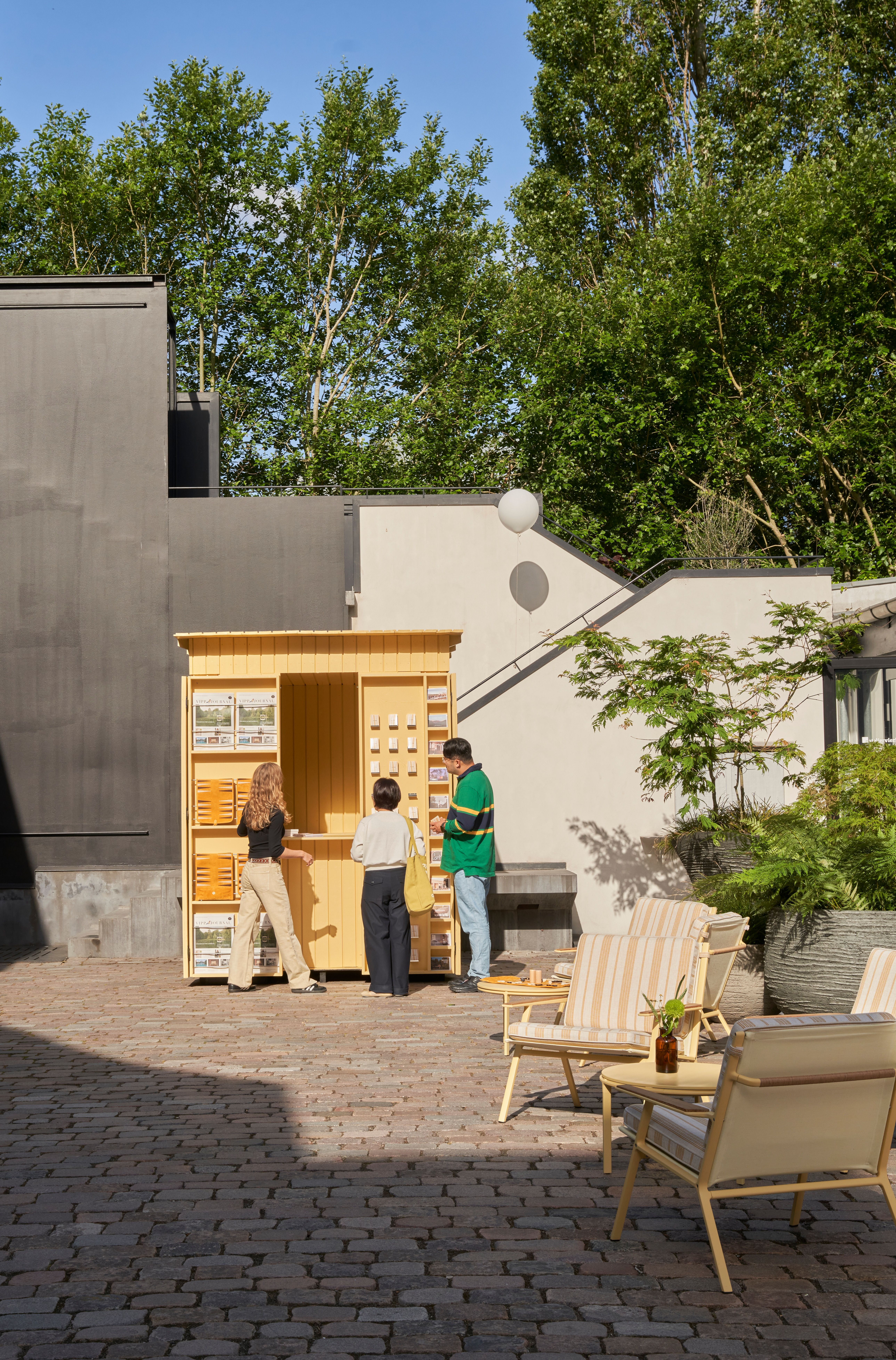 A playful installation comprising a chrome-clad, two-seat rocking chair, a butter yellow-painted kiosk, and a massive modular sofa with a square surface, butter yellow fabric, and chrome detailing.