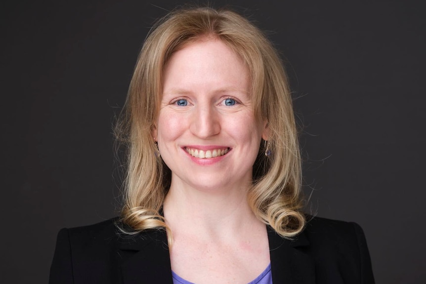 Woman with blonde hair smiles. She is wearing a purple shirt with a black blazer and is in front of a blank, grey screen.