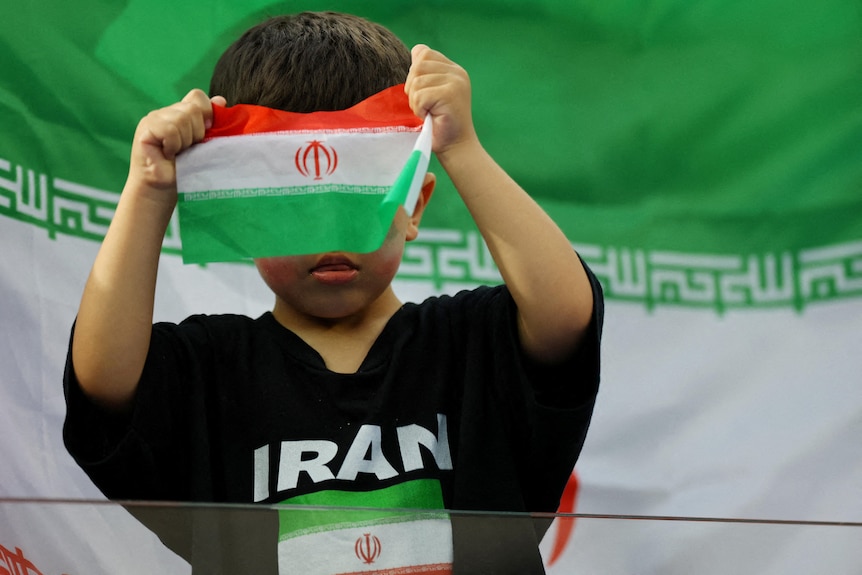 a small child holds a tiny islamic republic of iran flag in front of his face