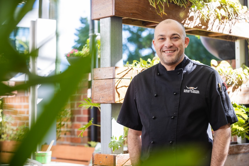A man wearing a chef's uniform with a "Norman Hotel" logo is smiling.