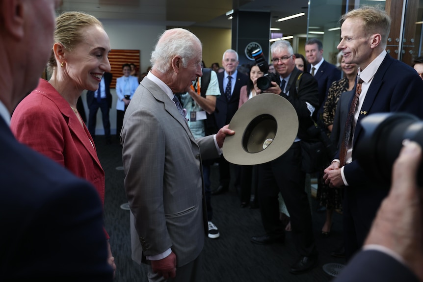 A group gathers around King Charles, smiling, as he looks down at an Akubra hat he has been gifted