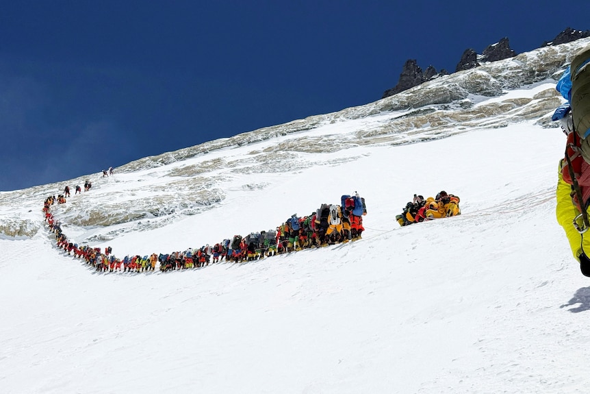 More than 100 climbers walk single file on a snowy mountain.