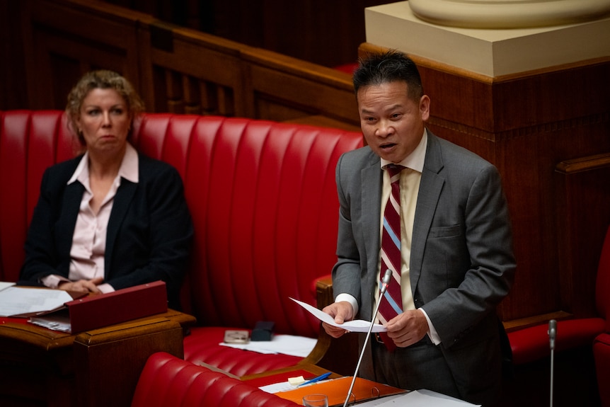A man with short black hair wearing a suit and striped tie speaks into a microphone in front of a red bench.