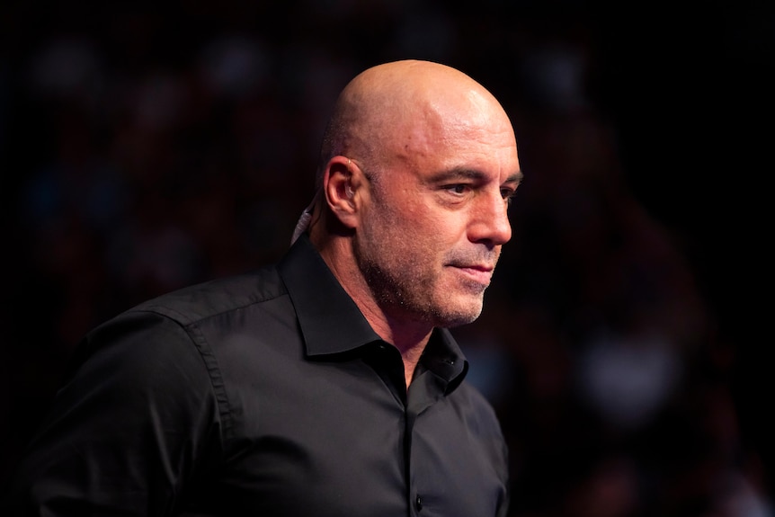 Joe Rogan wearing a dark shirt standing on a dark stage.