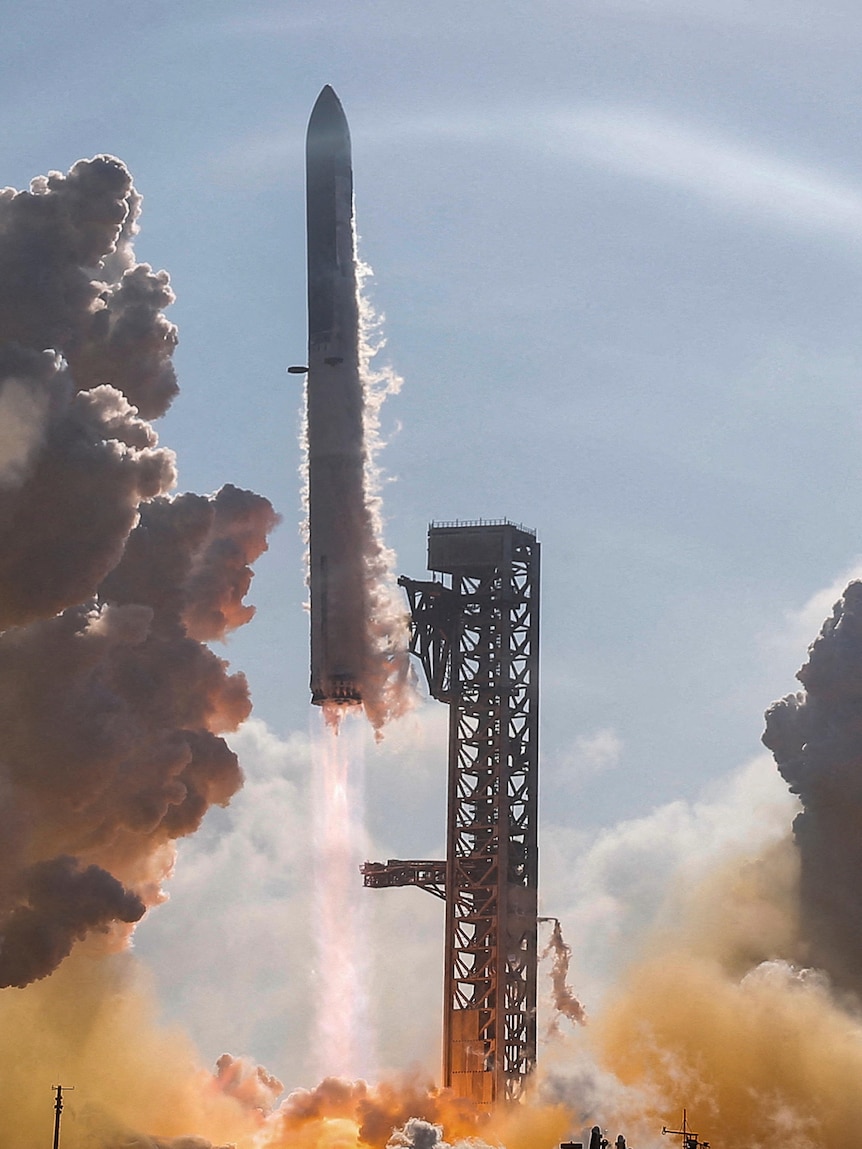 A white rocket launches into the sky on a clear day, with grey and yellow clouds billowing below.