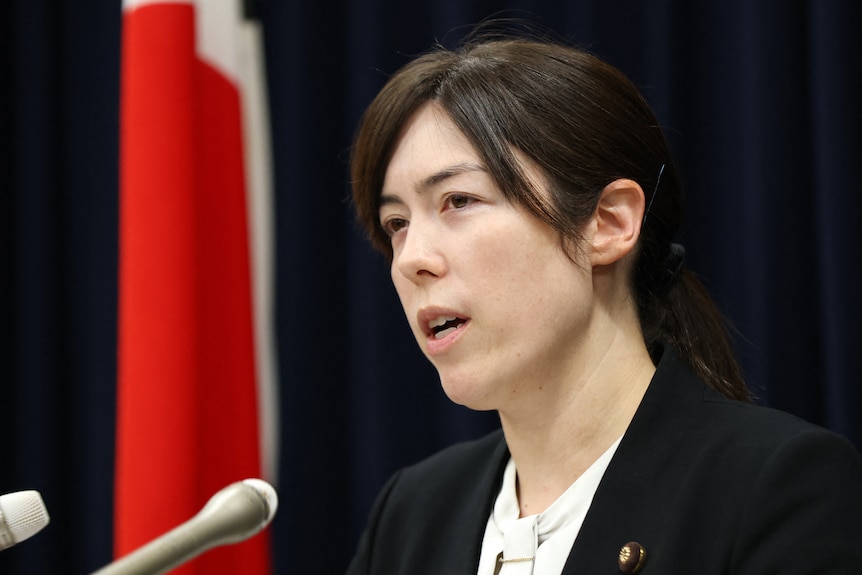 A woman with dark hair and in a dark jacket speaks at a microphone next to a Japanese flag.