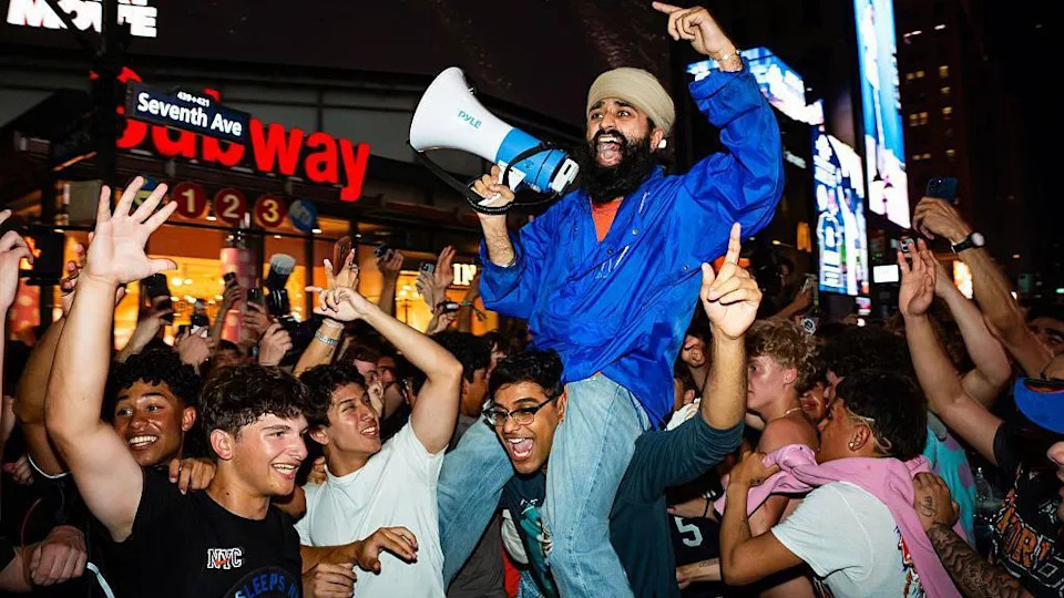 Crowds of fans cheering and celebrating, with one fan sitting on another's shoulders and yelling into a megaphone