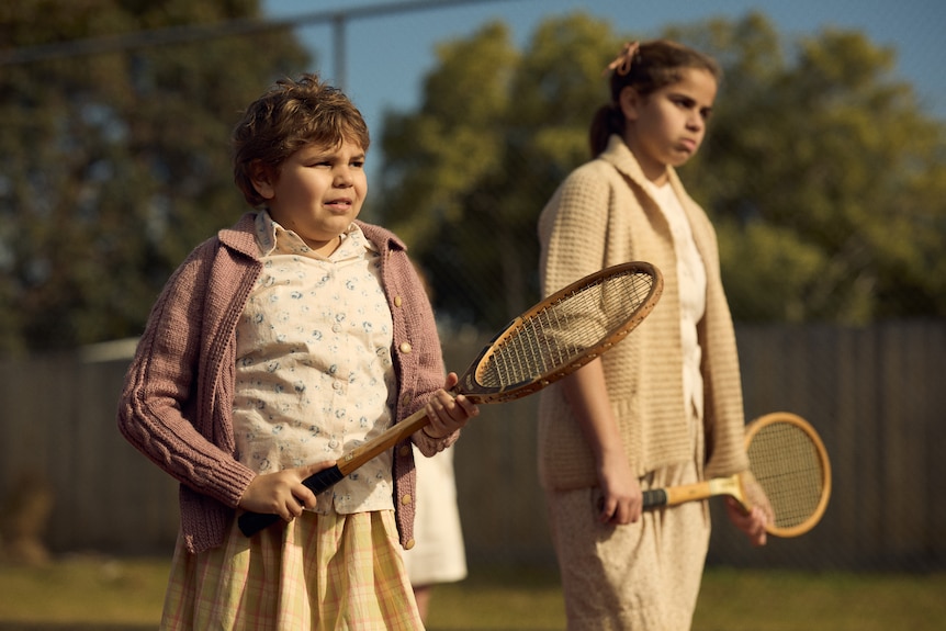 A young girl with cropped hair holds a wooden tennis racket and wears a knitted cardigan and a tennis skirt.