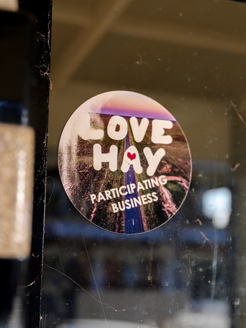 A sticker reading 'Love Hay participating business' stuck on a window with a reflection of a building in the glass.