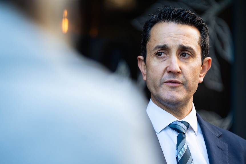 A man in a blue suit watching someone speak at a press conference.
