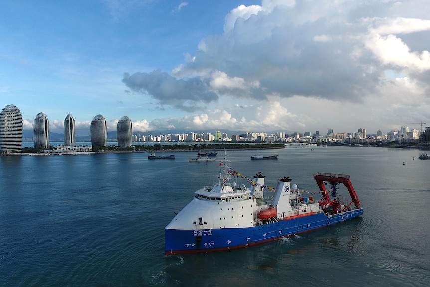 A large ship with white cabin at front, blue bottom and large flat back deck in port in front of large city.