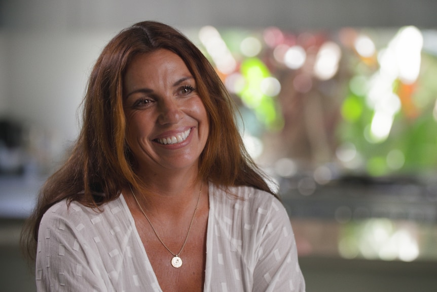 A woman with long brown hair wearing a white top sits in a kitchen and smiles.