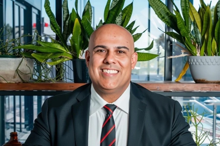 Man in a suit, red and black tie, sitting and smiling for a photograph.