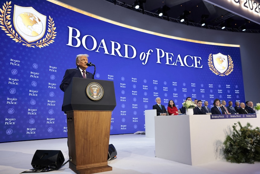 Man in suit standing behind a podium next to a group of people sitting down.
