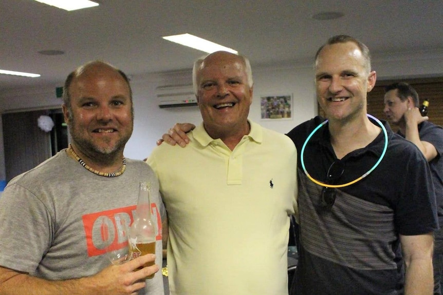 Three smiling men, one older, with their ams around each other at a venue.