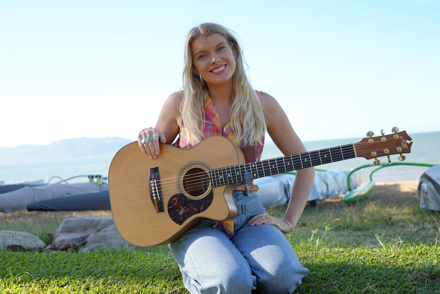 blonde woman with acoustic guitar sitting in front of beach
