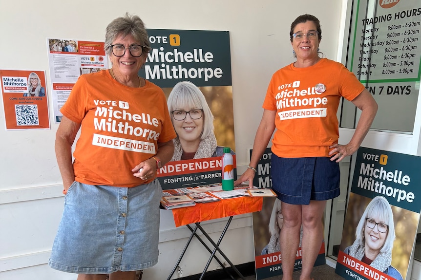Two women stand together wearing orange t-shirts with white writing, reading 'VOTE 1 Michelle Milthorpe Independent'.