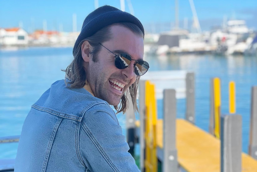 A man with long hair wearing a beanie and sunglasses looks ovcer his shoulder laughing and holding a rope at a dock.