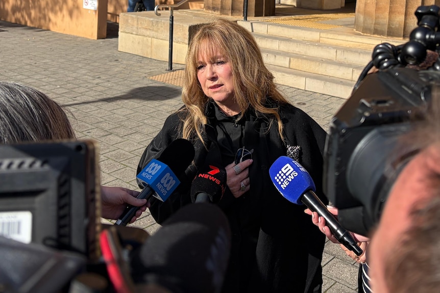 A blonde woman speaks to reporters holding microphones outside a stone building