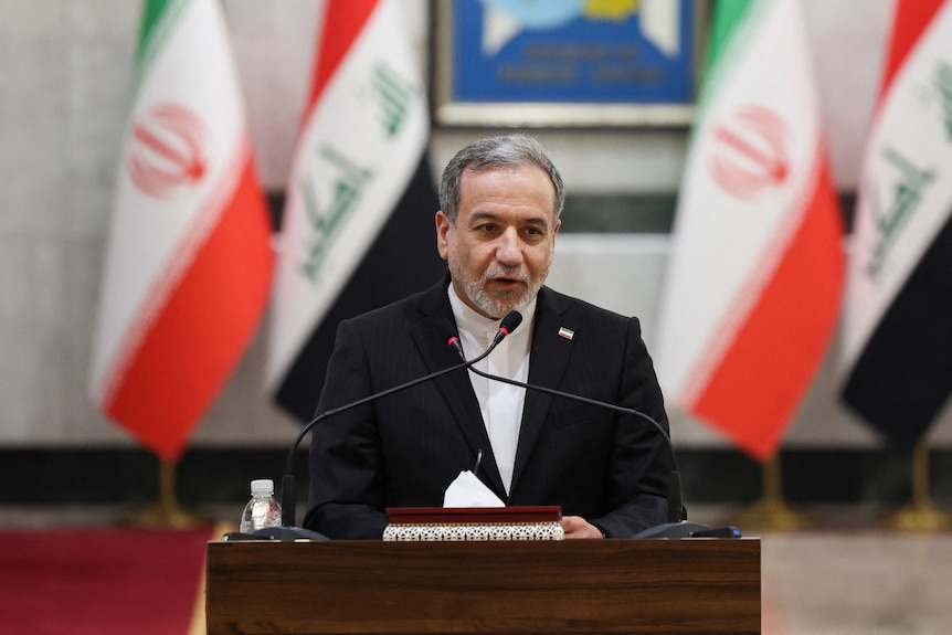 A man with gray, short hair, wearing a suit, stands behind a podium and speaks to a microphone, Iranian flags behind him