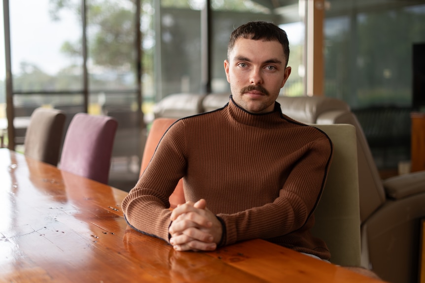 A man sits with fingers interlaced at a meeting table in front of large glass windows