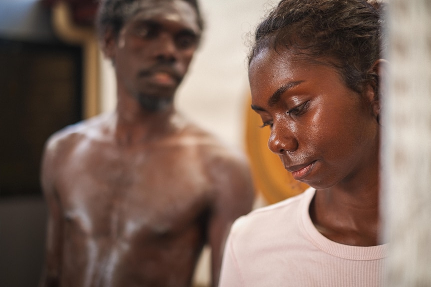 A young Indigenous woman looks over her left shoulder towards a shirtless man standing behind her.