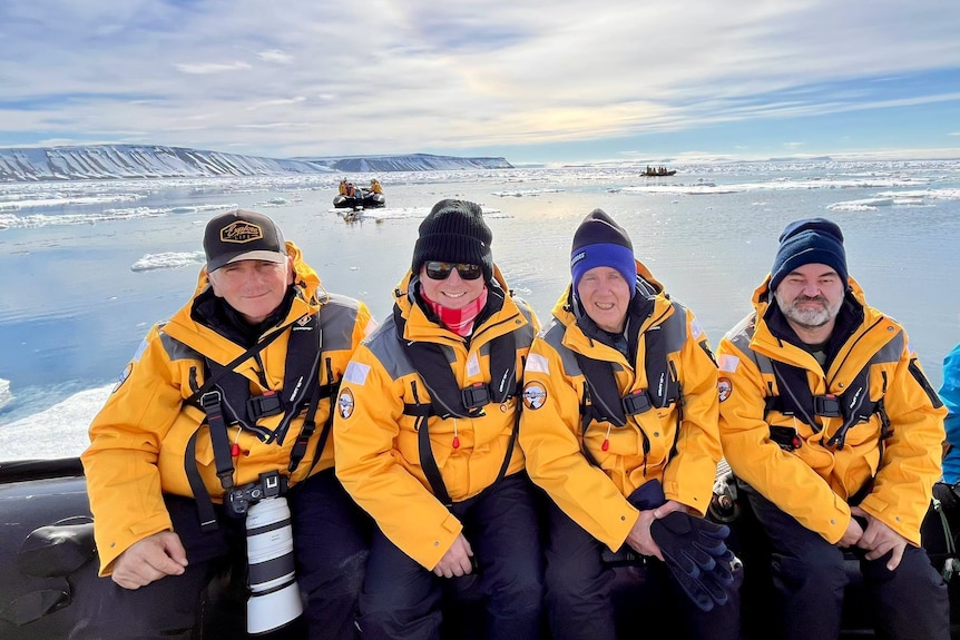 Four men in bright orange jackets and dark beanies sit in an inflatable boat in icy waters.