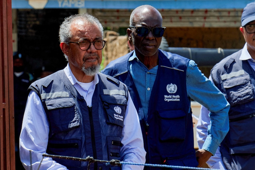 Three men standing wearing navy blue vests and blue shirts