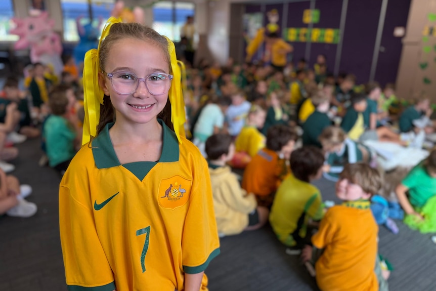 A young girl wearing glasses and yellow ribbons in her hair smiles at the camera with her fellow students sitting behind her.