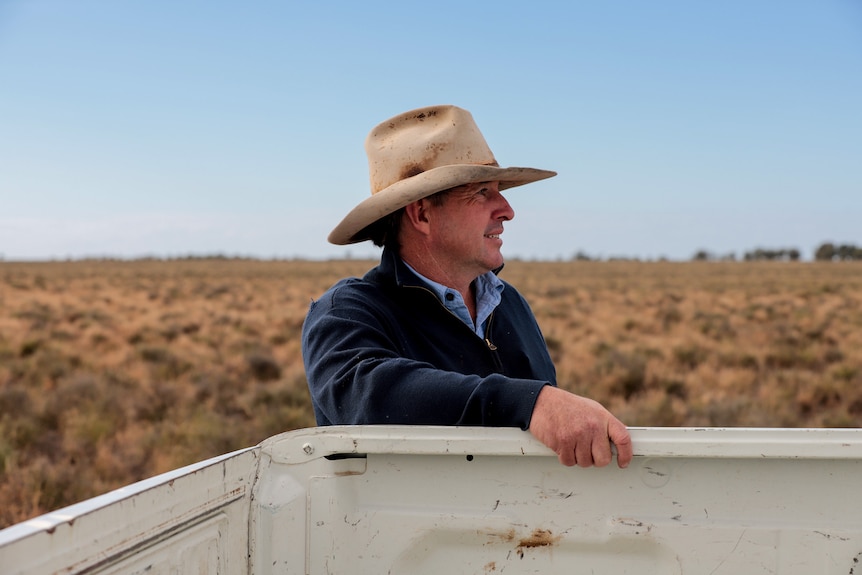 Man wearing weathered wide brim hat has hand on side of white ute tray with flat rural plains behind him