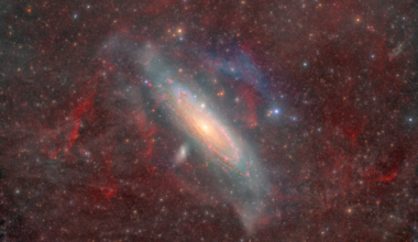 Wispy clouds of dust and gas in the Milky Way obscure the image. Milky Way stars are scattered across the image. Andromeda is a tight spiral of gas, dust, and stars that 
occupies the middle background. A couple smaller galaxies look like small bright clumps of stars near Andromeda.