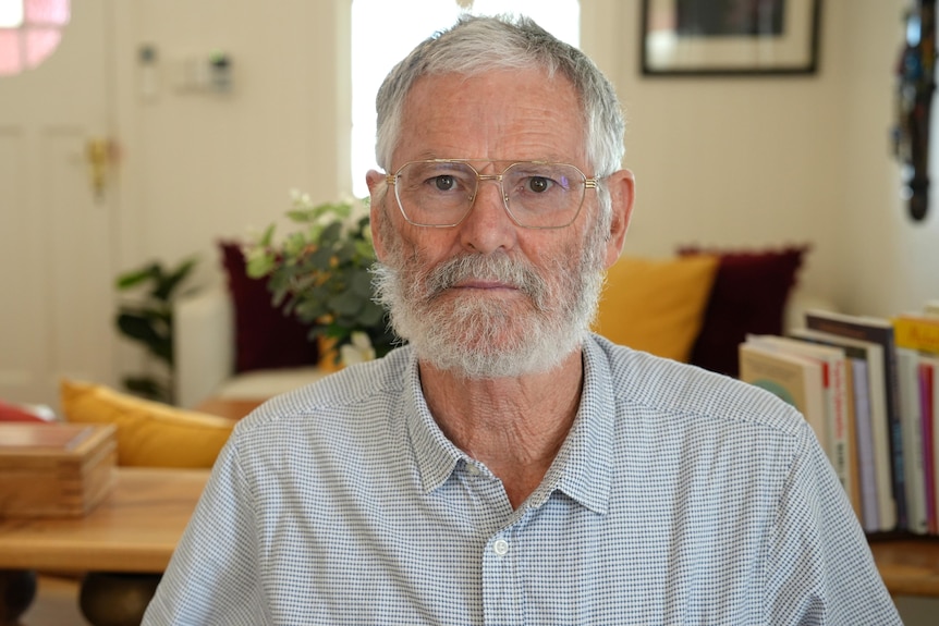 Photo of a man with a serious face and a beard, sitting in a room.
