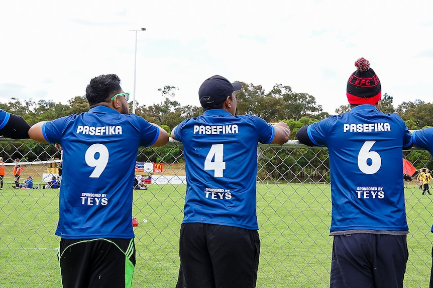 Seven men in blue soccer jerseys lean against a fence, watching a soccer match. Naracoorte, SA, November 2022.