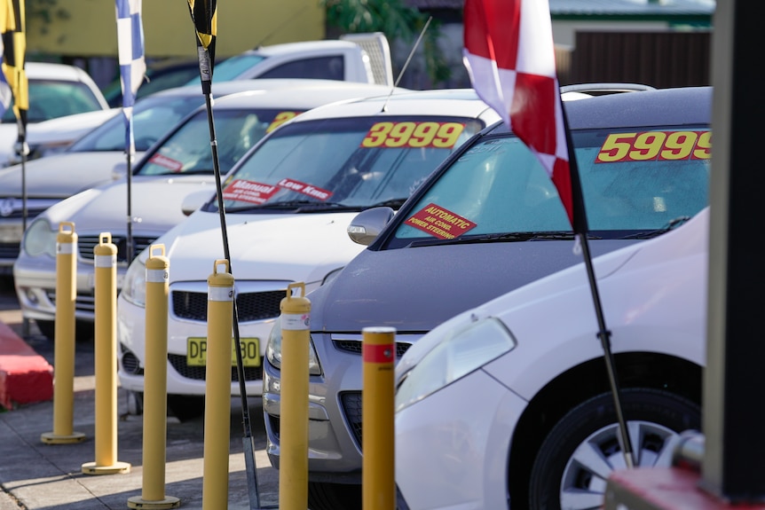 A row of cars in a car yard with red and yellow price stickers on them and flags across the front of the car park.