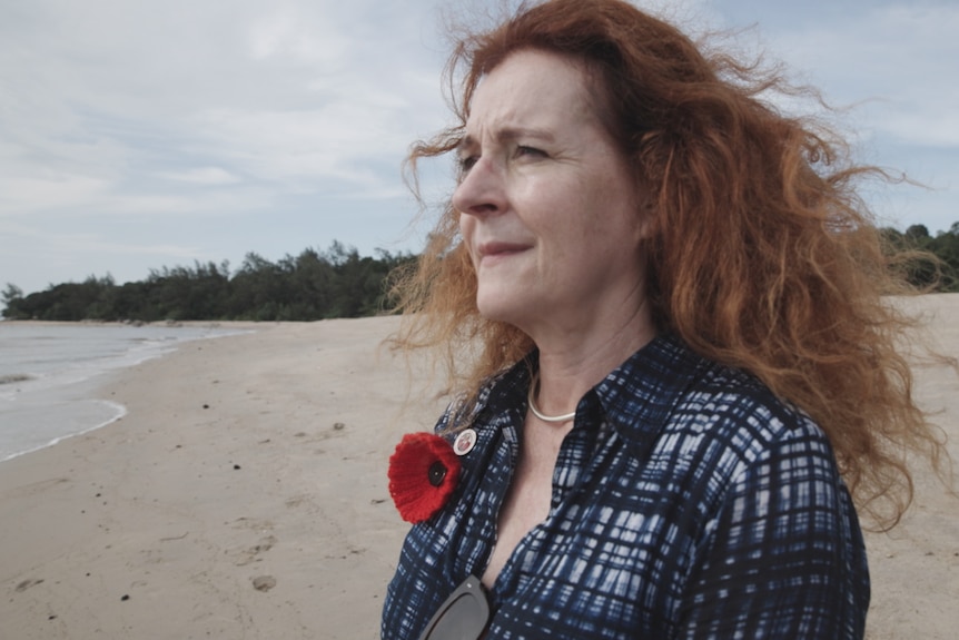 Woman with long hair blowing in wind stands on beach looking out to sea