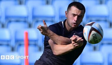 Charlie Chapman spins a pass out of his left hand sending the ball straight at the viewer. He watches it closely as it flies away from him. In the background there are empty light blue seats which are blurred and the top part of a red training stick.