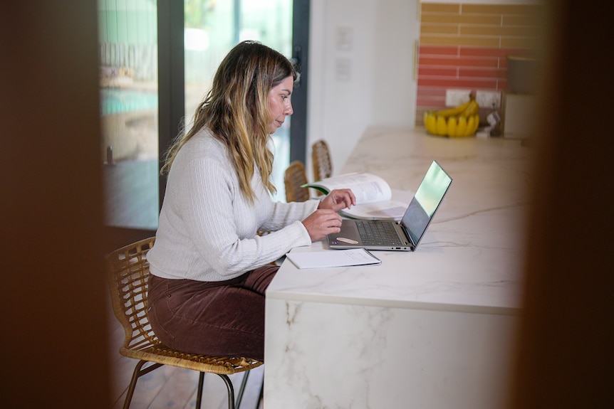 A woman in a white top and brown pants sits at her kitchen bench and looks at a laptop.