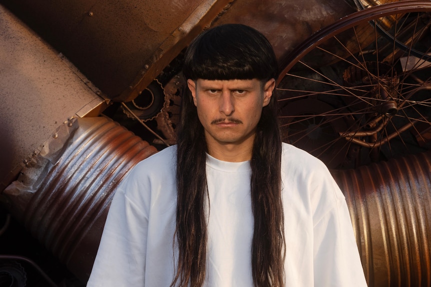 man with moustache and long-dark haired mullet stands stony faced in front of scrap heap