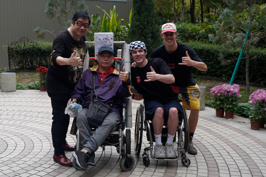 Two men in wheelchairs next to each other and two people standing either side of them with their thumbs up.
