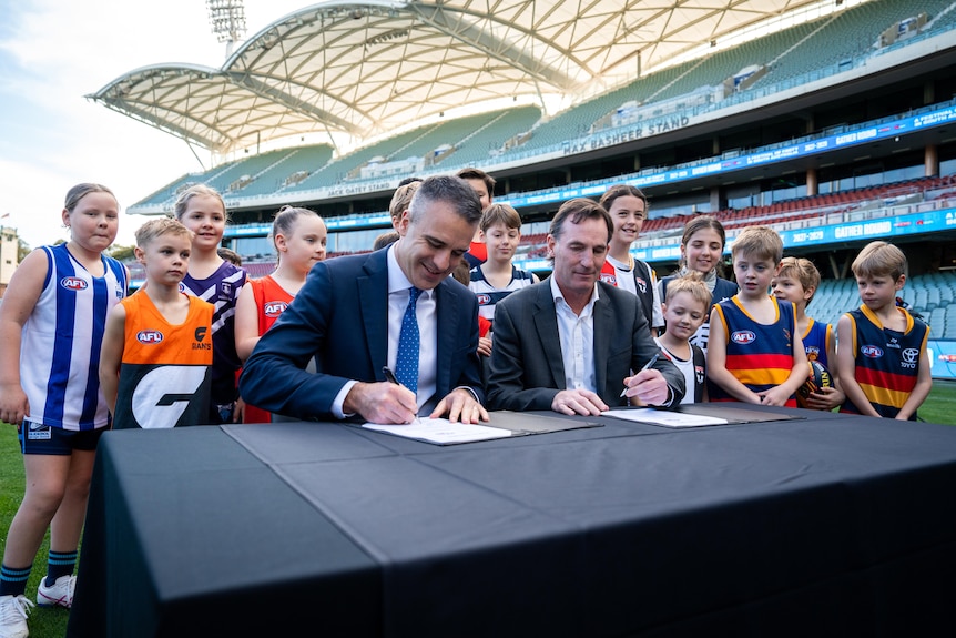 Two men sign papers on a black table while children wearing AFL jumpers look on within a stadium