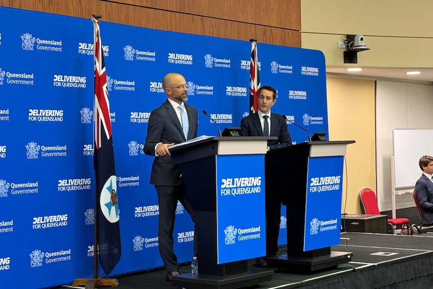 Two men in suits standing at podiums with flags on either side.