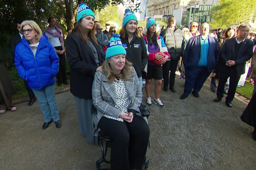 Two women wearing blue beanies stand behind a woman who is seated, wearing the same beanie, near a crowd of people.