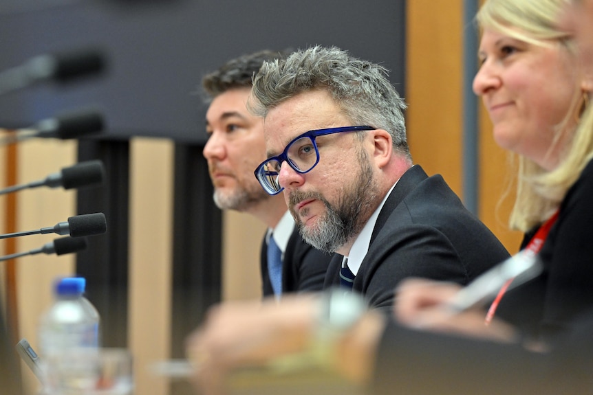 Justin with grey hair and glasses sits at a Senate estimates hearing, looks in front, pic taken from side. Man and woman beside.
