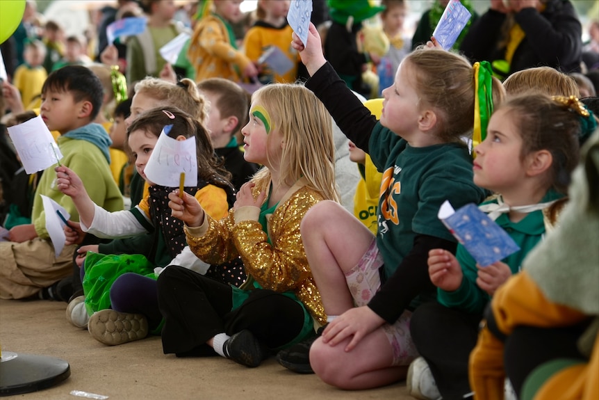 A long row of young school children wearing green and gold wave handmade Australian flags on paddlepop sticks.
