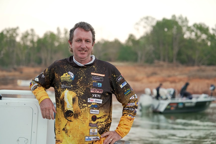 White man with brown / gray hair, wearing long sleeve yellow and black hoodie, smiling, standing on boat in river.