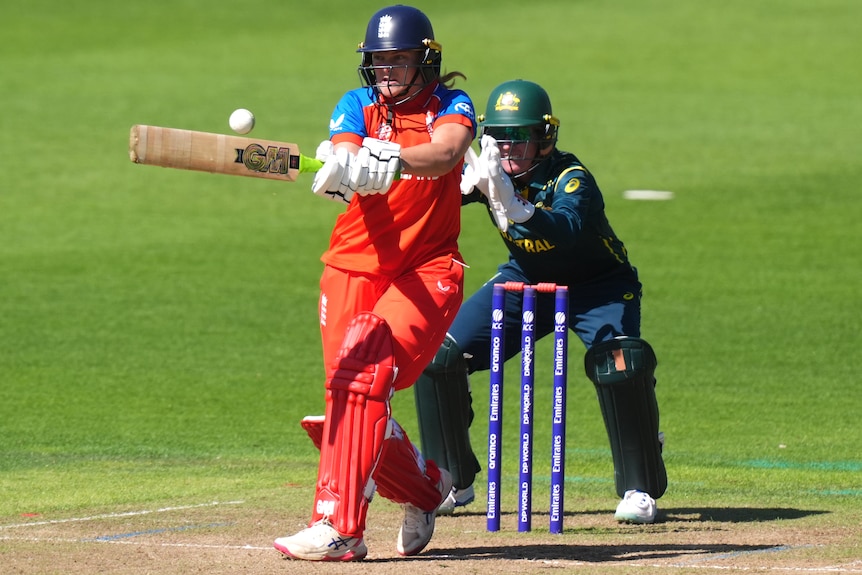 Dani Gibson swings her bat to hit the ball. Behind her, the Australian wicketkeeper has her hands out, ready.